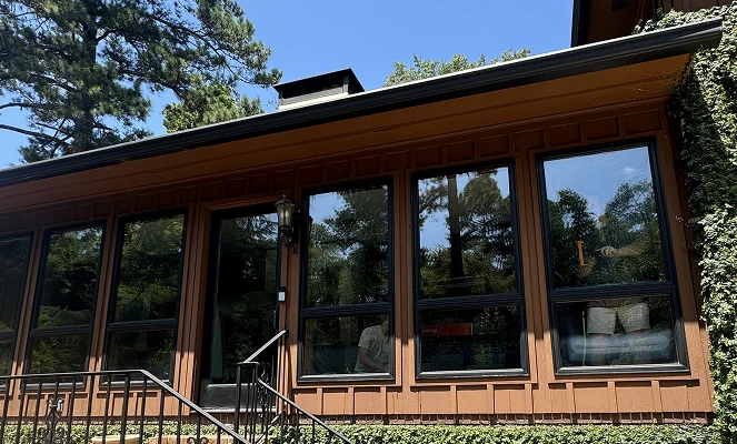 Exterior of a brown wooden home with large reflective windows showing the surrounding trees and sky. The windows appear tinted, offering privacy and UV protection while blending with the home’s natural aesthetic.