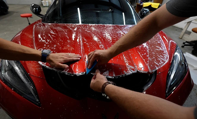 Close-up of two technicians applying Paint Protection Film to the hood of a red sports car, using blue squeegee tools to smooth out the wet film. Water and slip solution are visible across the surface, aiding in precise placement.