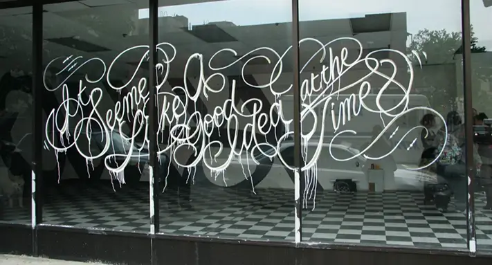 Large storefront windows with white graffiti-style script reading “It seemed like a good idea at the time,” written in decorative cursive with paint dripping down.