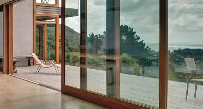 Modern home with large floor-to-ceiling sliding glass doors framed in wood, offering a scenic view of trees, hills, and the ocean beyond a spacious deck.