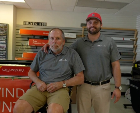 Two men wearing matching gray polos with a window tinting logo pose inside a workshop. One man is seated while the other stands beside him with an arm around his shoulder. Behind them are rolls of film and signage for Paint Protection Film and Window Tint.