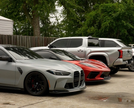 Three high-end vehicles—a matte gray BMW, a red Chevrolet Corvette, and a silver Toyota Tundra—are parked in a lot outside, surrounded by a wooden fence and trees. All vehicles appear to have tinted windows and well-maintained exteriors.