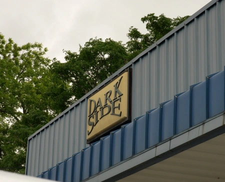 Exterior sign for "DARK SIDE" mounted on a blue metal-paneled building with trees and a cloudy sky in the background. The sign has a beige background with bold black lettering.