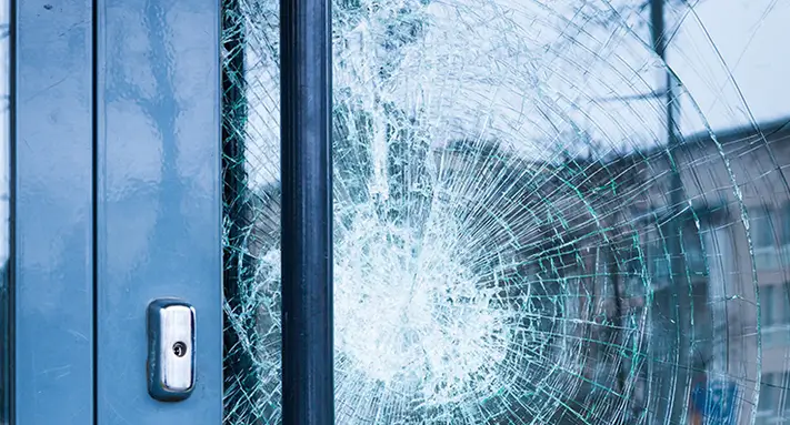 Close-up of a glass door with a large spiderweb-like crack, showing impact damage next to a metal handle and lock.