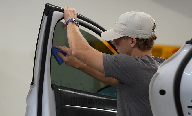 A technician in a light cap and gray shirt applies window tint film to the interior of a white vehicle’s door using a blue squeegee tool. The window is wet for proper film positioning and adherence.