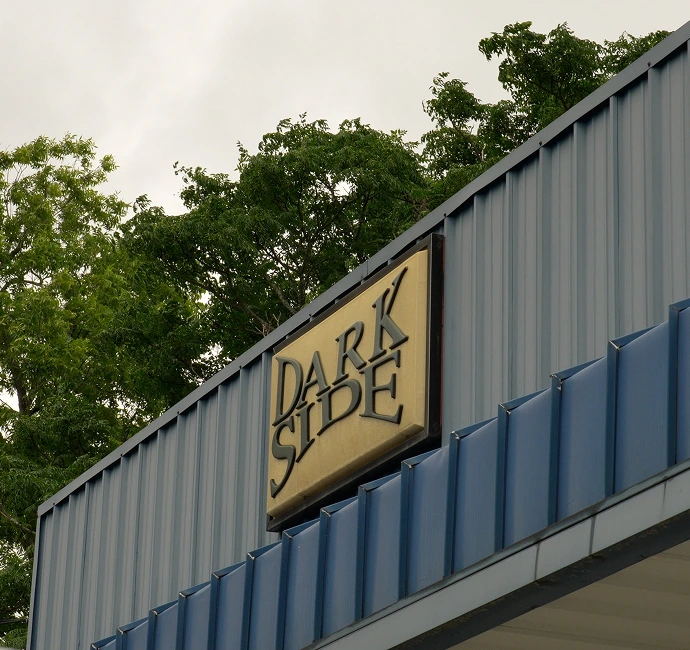 Outdoor sign reading "DARK SIDE" in bold, serif letters mounted on the exterior of a blue metal-paneled building. The sign has a beige background and is angled upward, with green trees visible behind the structure.