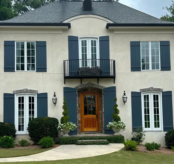 Elegant two-story home with cream-colored stucco walls, dark blue shutters, arched wooden front door with stone trim, and a small balcony above the entrance.