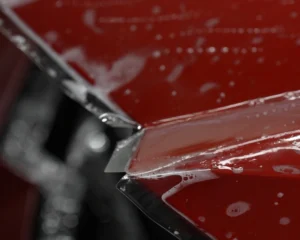 Close-up of a transparent paint protection film being applied to the edge of a glossy red car panel, with visible soapy water and air bubbles during installation.