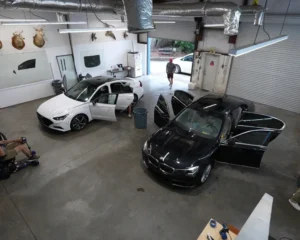 Interior of an auto detailing or tint shop with a white sedan and a black BMW parked side by side, both with doors open while technicians work on them.
