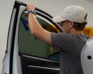 A man wearing a beige cap uses a blue squeegee to apply window tint film to the inside of a car door window.