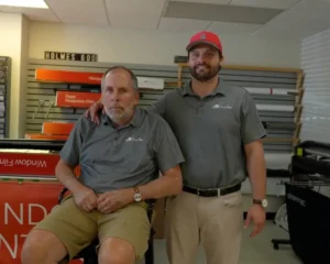 Two men wearing matching gray "LLumar SelectPro" shirts pose together inside a window tint and film shop, with one seated and the other standing beside him, resting a hand on his shoulder.