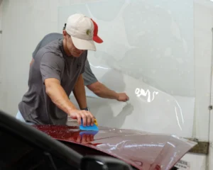 A man in a beige cap and gray shirt smooths out a paint protection film on a red car hood with a blue squeegee, while another person in the background prepares more film.