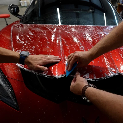 Two technicians apply Paint Protection Film to the wet, soapy hood of a red sports car using a blue squeegee and precise hand placement. Reflections from overhead lights are visible on the glossy surface.