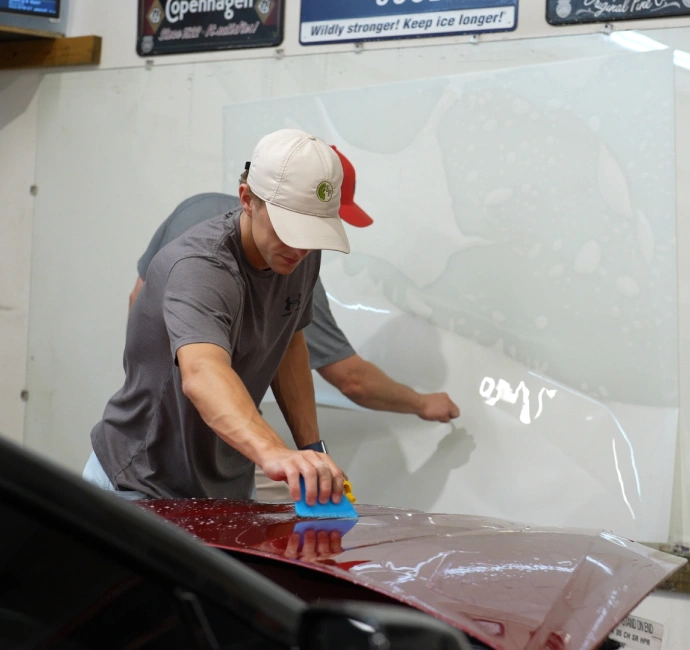 A man in a gray shirt and beige cap applies a transparent paint protection film to the hood of a red car using a blue squeegee, while another person handles film in the background.