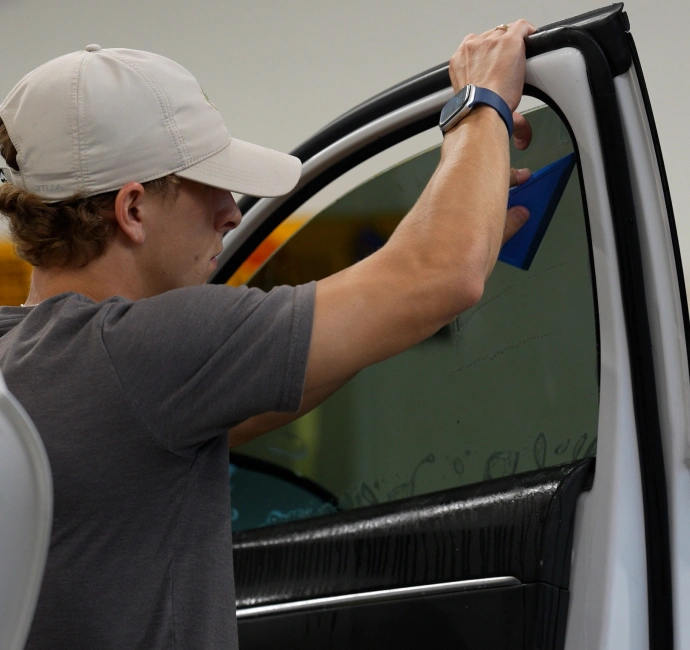 A man wearing a gray shirt and beige cap uses a blue squeegee tool to apply window tint film to a car door’s glass.