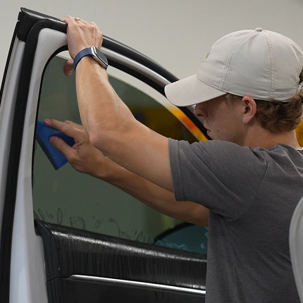 Technician in a gray shirt and beige cap uses a blue squeegee to apply window tint film to the interior of a white vehicle's door glass. The film is being smoothed out over a wet surface for optimal adhesion.