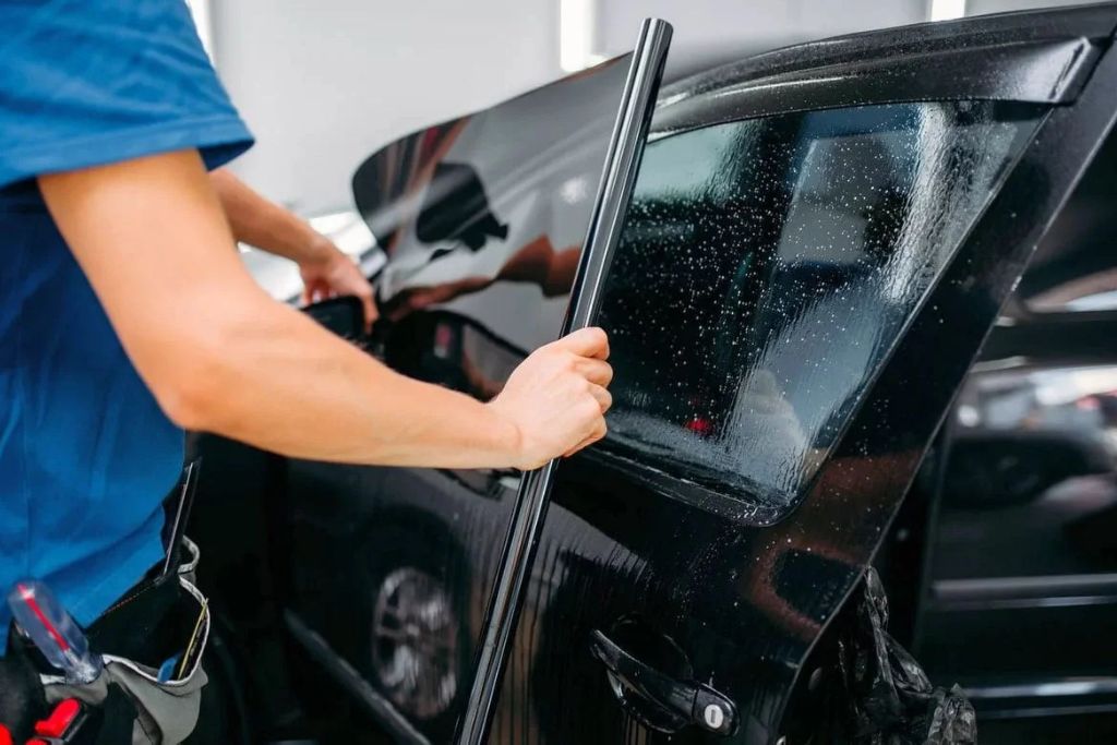 A technician carefully installs a dark window tint film onto a car door window, with water droplets visible on the glass to aid application. This step highlights what is window tinting by showing the process of enhancing privacy, reducing glare, and blocking heat with specialized film.