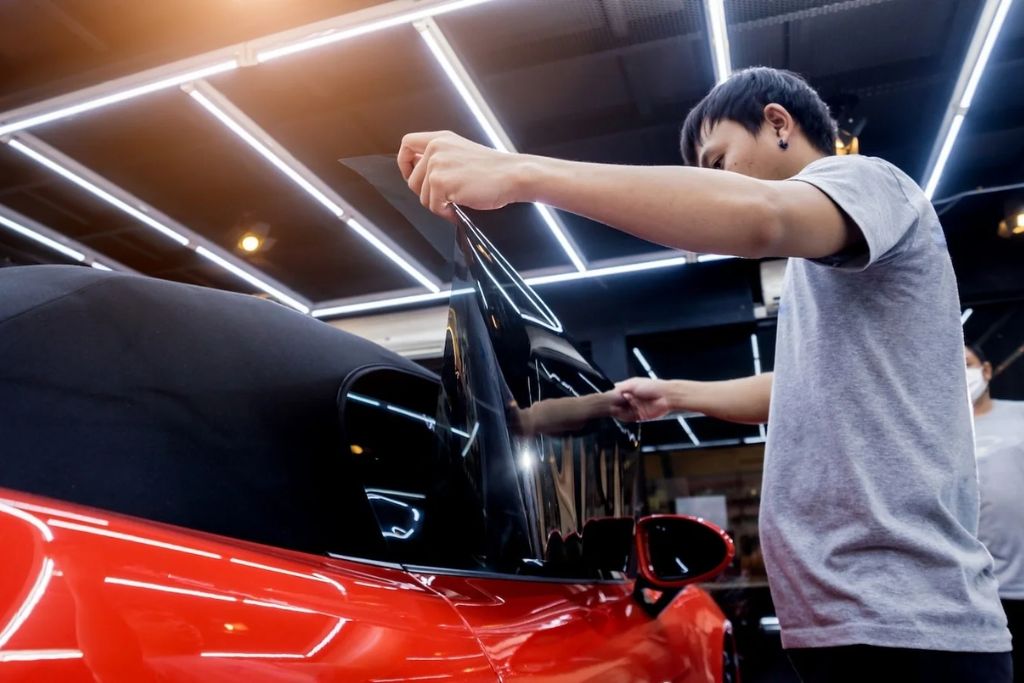 A technician installs dark window tint film onto a red car’s side window under bright workshop lighting, showcasing the application of ceramic tint for improved UV protection and heat rejection. This image demonstrates what is window tinting by highlighting the use of high-performance film to enhance comfort, durability, and style.