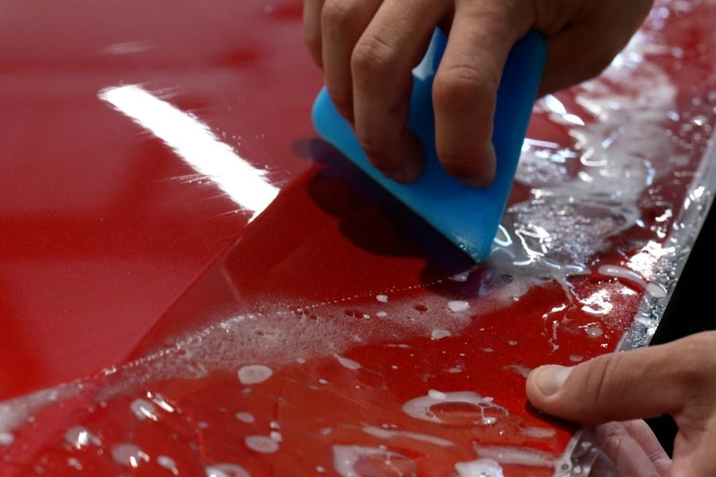 Dark Side expert smooths paint protection film on a red car using a blue squeegee.