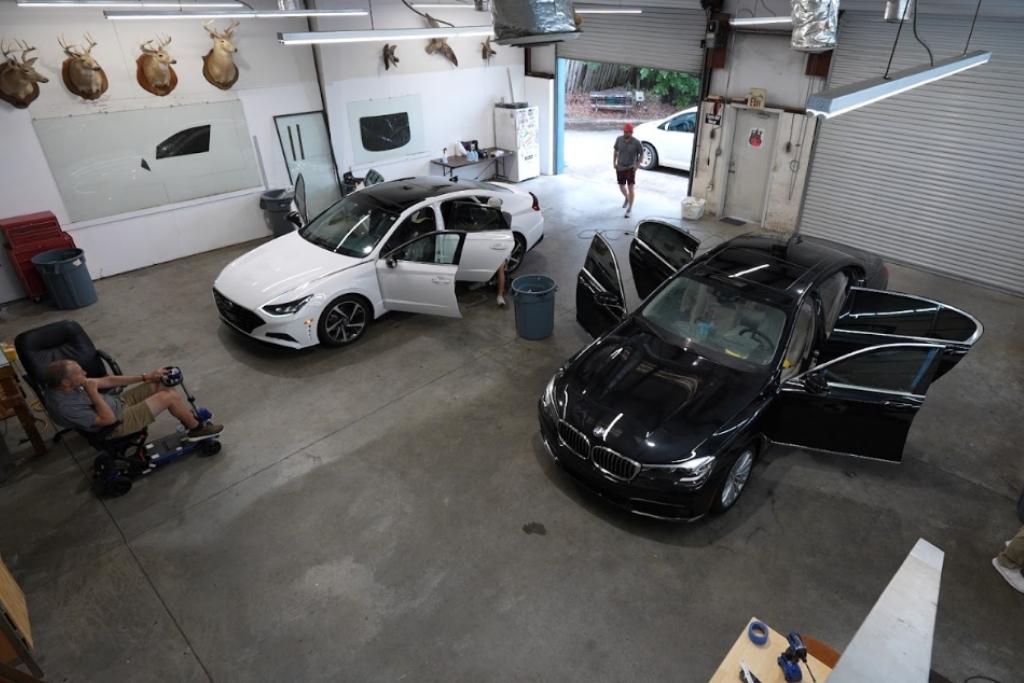 Interior of an automotive tint shop with two cars being worked on, technicians installing window film, and a customer seated nearby.