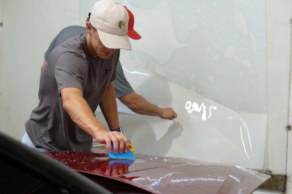 Two technicians applying paint protection film on a red vehicle in a professional shop.