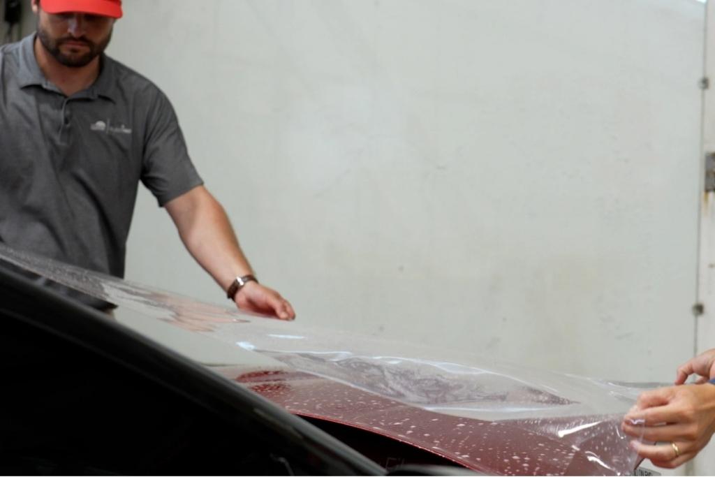 Technicians applying paint protection film to the hood of a red car during professional installation.