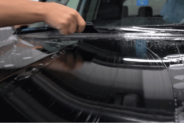 Hand smoothing paint protection film on a glossy black car hood.