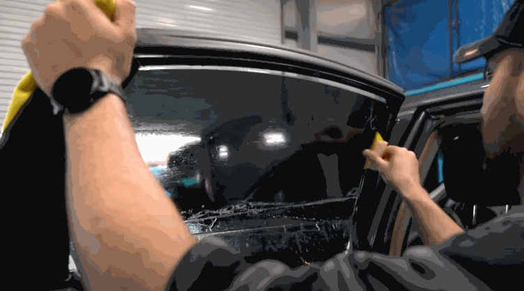 Installer smoothing out window tint film on a vehicle’s door glass using a yellow squeegee and cloth.