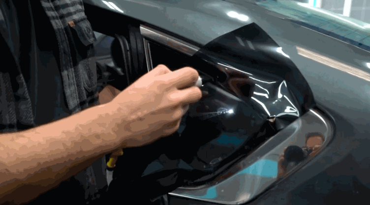 Technician carefully positioning dark window tint film on the corner of a car window while using a spray bottle.