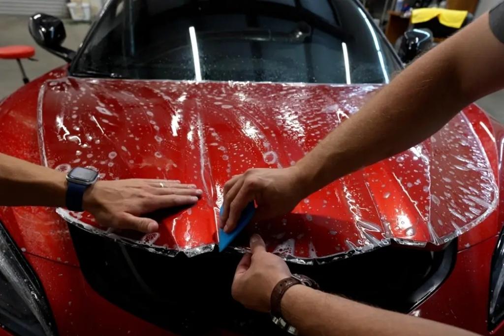 Technicians applying clear paint protection film to the hood of a red vehicle using a squeegee during installation.
