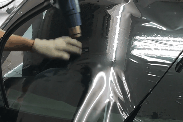 Technician using a heat gun to apply and shape window tint film on a curved car window surface
