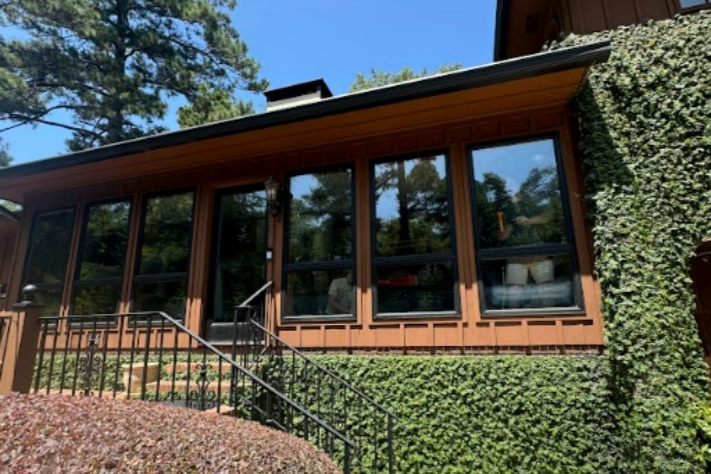 Exterior of a wood-sided home with large dark-tinted windows surrounded by trees and greenery.