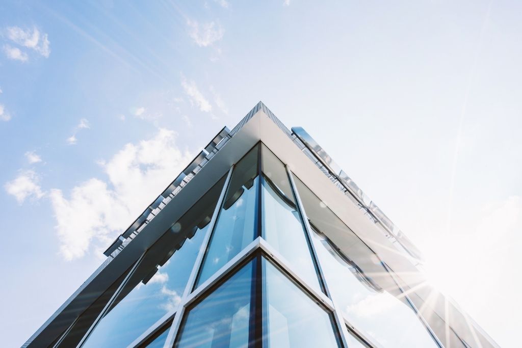 Low-angle view of a modern commercial building with large glass windows reflecting the sky and bright summer sunlight.
